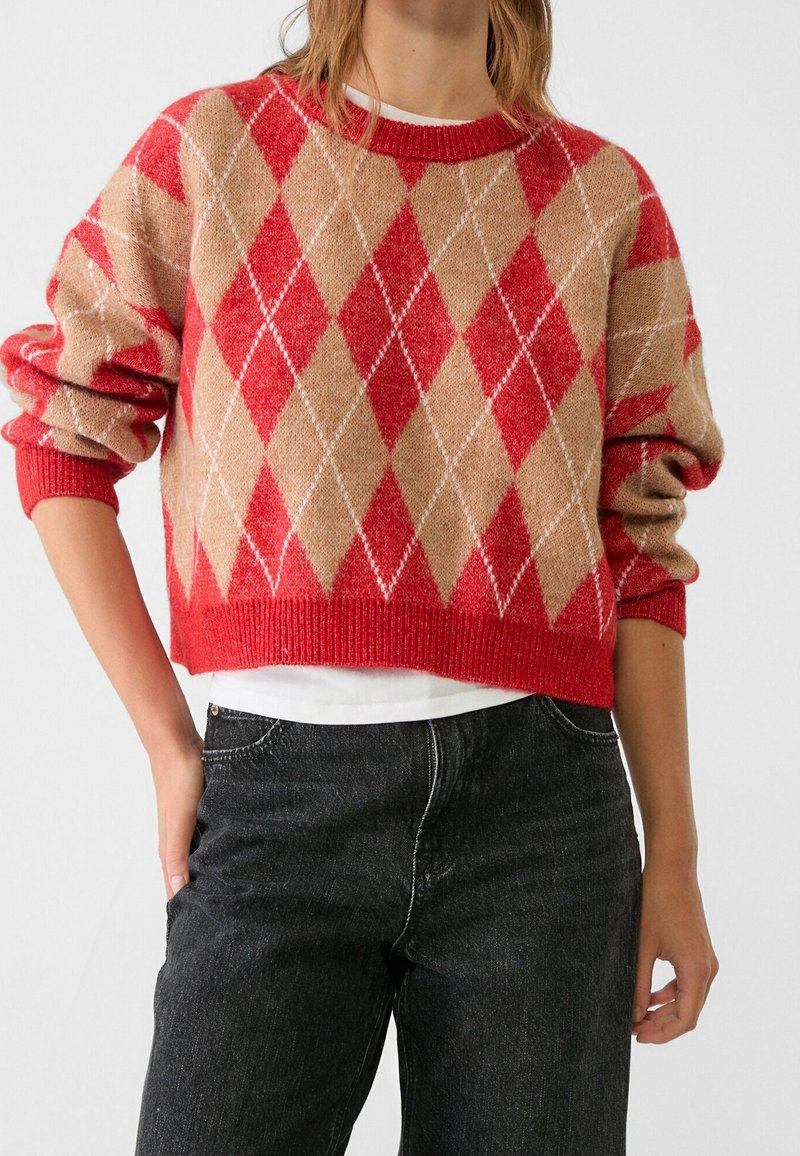 Personne portant un pull à motif argyle rouge et beige avec les manches retroussées sur une chemise blanche et un jean noir.