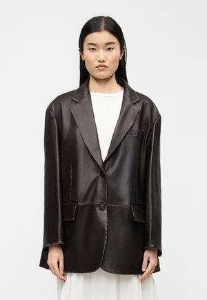 Jonge vrouw met lang zwart haar draagt een oversized donkerbruine leren blazer en een witte jurk, staand tegen een effen lichte achtergrond.