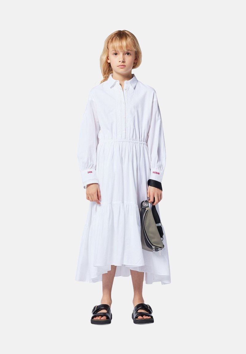 Witte jurk met lange mouwen en een gelaagde zoom, knopen aan de voorkant en een elastische taille. Zwarte sandalen en een grijze tas maken de look compleet. Rode tekst op de mouwen.