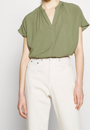 Femme portant un chemisier vert olive à manches courtes et col en V, rentré dans un pantalon blanc taille haute, sur un fond clair uni.