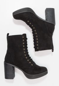 Bottes cheville en daim noir avec un talon chunky, des œillets dorés et un laçage avant. Caractérisées par un talon texturé et un bout rond lisse.