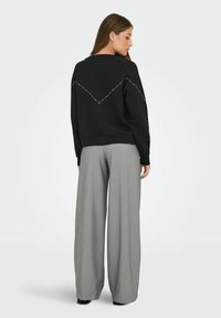 Femme de dos portant un sweat-shirt noir avec un motif en chevrons à pois dans le dos et un large pantalon gris devant un fond blanc.