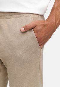 Pantalons beiges en tissu texturé. Présentent une poche latérale avec une couture visible et une main reposant sur la poche, mettant en valeur le matériau.