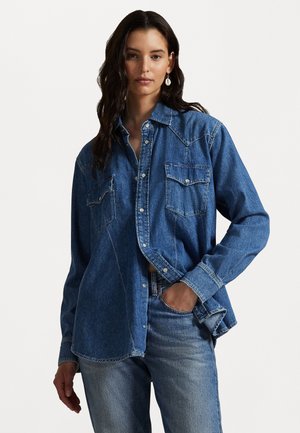 Camisa de ganga azul com mangas longas, dois bolsos no peito, gola pontuda e fecho frontal com botões. Usada com jeans azul claro.