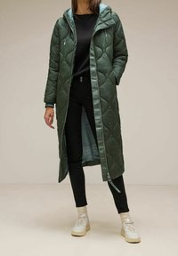 Abrigo largo acolchado de color verde oliva con diseño acolchado, capucha y cremallera frontal; combinado con pantalones negros y zapatillas ligeras.