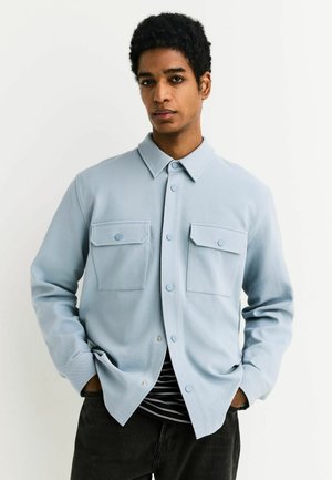 Young man with short curly hair wearing a light blue buttoned shirt jacket with two chest pockets and black pants.