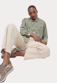 Giubbotto di jeans verde con tasche frontali, pantaloni lunghi beige e sneaker color marrone chiaro con dettagli più scuri; modello seduto con una gamba incrociata.
