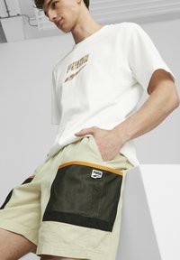 T-shirt bianco in cotone con logo Puma dorato, abbinato a pantaloni corti verde chiaro con tasche in rete nera e dettagli arancioni.