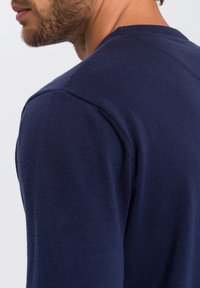 Granatowy sweter wykonany z dżerseju, z okrągłym dekoltem i ściągaczami na rękawach. Zbliżenie na ramię i okolice dekoltu.