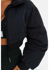 Vista recortada de una persona que lleva una chaqueta acolchada negra sobre un top de punto cortado verde oscuro y pantalones.