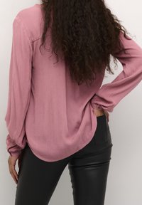 Blouse rose clair à manches longues en tissu texturé, présentant une coupe décontractée avec un ourlet arrondi et des poignets boutonnés, associée à un pantalon noir.