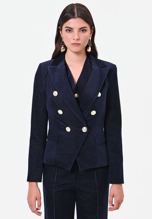 Femme aux cheveux raides et foncés porte un blazer bleu marine à double boutonnage avec des boutons argentés et un pantalon assorti, debout devant un fond uni.