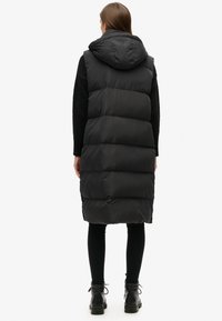 Schwarzte gepolsterte Weste mit Kapuze, knielang, mit einem gesteppten Design und strukturiertem Stoff. Getragen über schwarzen Leggings und Ankle Boots.