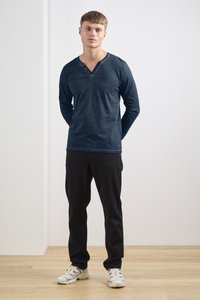 Langärmliges navyfarbenes V-Ausschnitt-Shirt mit Knopf-Details, kombiniert mit schwarzen Hosen und hellen Sneakers, stehend auf einem Holzboden.
