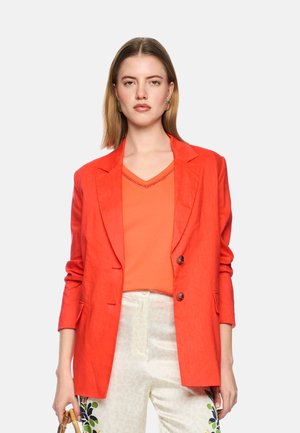 Woman with straight blond hair wears bright orange blazer and top, white pants with floral print, holding a small wooden handbag.