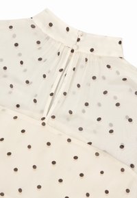 Kreemi-värvi blouse, millel on läbipaistev kangas, millel on laiali jaotatud pruunid padjakesed, kõrge kaelus ja nööbiga kinnitamine seljaosas.