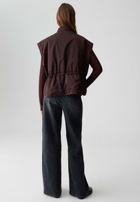 Bruine mouwloze jas met hoge kraag, met een strakke taille, gedragen over een aansluitend long-sleeve shirt en gecombineerd met wijde zwarte jeans.