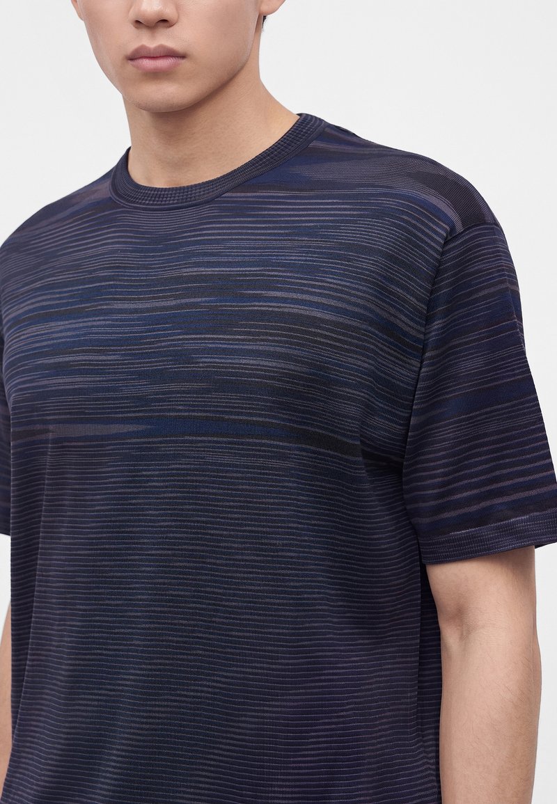 Persona joven con una camiseta de manga corta azul oscuro y negra con finas rayas horizontales y un escote redondo.