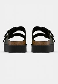 Μαύρα σανδάλια πλατφόρμα με δύο λουριά με πόρπες, πάτο από φελλό και καουτσούκ σόλα. Ο ανοιχτός σχεδιασμός στη φτέρνα αναδεικνύει την αντιθετικότητα της υφής μεταξύ των υλικών.