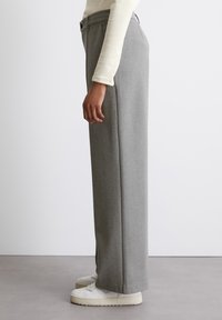 Pantalons large gris clair en tissu doux avec une texture lisse, présentant des plis sur le devant et des poches latérales. Associés à des baskets blanches.