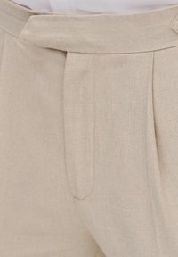 Jasnobeżowe lniane spodnie o plisowanym fasonie, gładkiej fakturze i dopasowanym pasku w talii. Brak widocznych kieszeni.