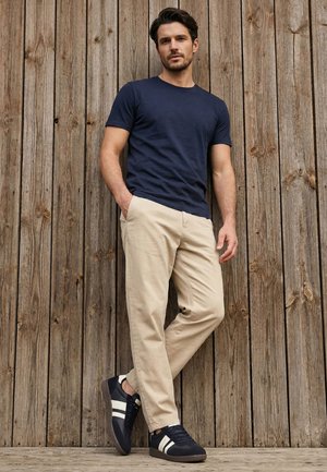 Muž opírající se o dřevěnou stěnu, má na sobě tmavě modré tričko, béžové kalhoty a černé tenisky s bílými pruhy.