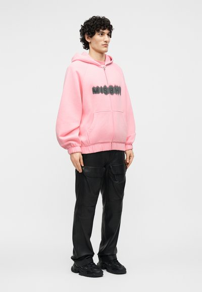 Jeune homme portant un sweat à capuche zippé rose avec un texte noir et un pantalon cargo noir, debout devant un fond clair uni.
