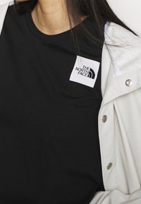 Mujer con una camiseta negra de The North Face y una chaqueta clara de botones beige sobre el hombro, con el cabello largo y oscuro.