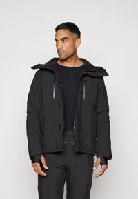 Schwarze isolierte Jacke mit Kapuze, ausgestattet mit Reißverschlusstaschen, verstellbaren Bündchen und strukturierter Stoffoberfläche. Trägt man über einer schwarzen Basislage.