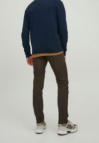 Dunkelblauer Pullover, hellbraunes Unterhemd, schmal geschnittene braune Hose und helle Sneakers, von hinten betrachtet, um die Details des Outfits zu zeigen.