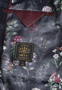Blomstermønsteret stof i nuancer af mørkeblå og grøn med burgunder akcenter. Har et mærke for "NEXT Tailored Fit," størrelse 42R.
