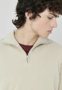 Pull en beige zippé avec un col montant, en tissu tricoté texturé et une fermeture éclair en métal. Porté par-dessus un t-shirt à col rond bordeaux.
