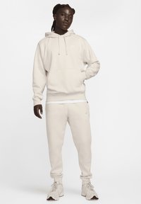 Gai smilškrāsas Nike kapuce ar lielo kabatu un saites, kombinēta ar atbilstošām sporta biksēm. Tekstūrēta auduma ar smalku logotipu uz krūtīm un gurniem.