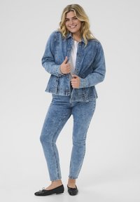 Chaqueta de denim con un lavado azul claro y cierre de botones, combinada con unos pantalones vaqueros a juego en un tono similar. Unos zapatos planos negros completan el atuendo.