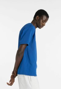 Blå bomuld T-shirt med rund halsudskæring og korte ærmer, vist fra siden mod en hvid baggrund, kombineret med lysegrå shorts.