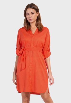 Mujer con vestido camisa naranja de manga larga y largo hasta la rodilla, con botones delanteros y cinturón atado a la cintura, sobre un fondo liso y claro.