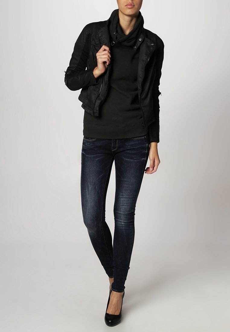 Chaqueta de piel sintética negra con detalles en los hombros, suéter de cuello alto verde, jeans ajustados azul oscuro y zapatos de tacón altos negros.