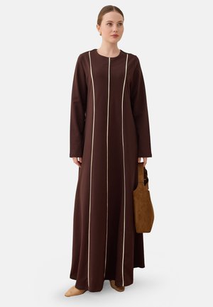 Vrouw draagt een lange bruine jurk met witte verticale strepen, houdt een bruine suède shopper vast, staand tegen een effen achtergrond.