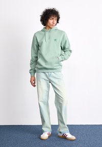 Mintgroene hoodie met kappmouw en voorzak, gecombineerd met lichtgewicht spijkerbroek. Model draagt witte schoenen met rode strepen.