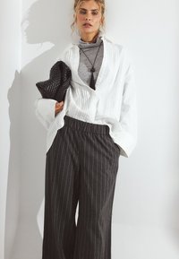 Bela srajca z gumbi in vertikalnimi naborki, sivi pulover s kolenom spodaj, črna pletena torbica, sivo-pasaste široke hlače.