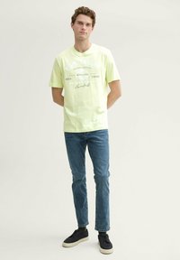 Lys grøn bomulds T-shirt med hvidt trykt logo og tekst, parret med blå jeans og sorte sneakers. Simpelt, afslappet design.