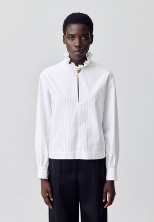 Modèle portant un chemisier blanc à rayures avec un col à volants et un fermoir doré, associé à un pantalon noir, sur un fond neutre.