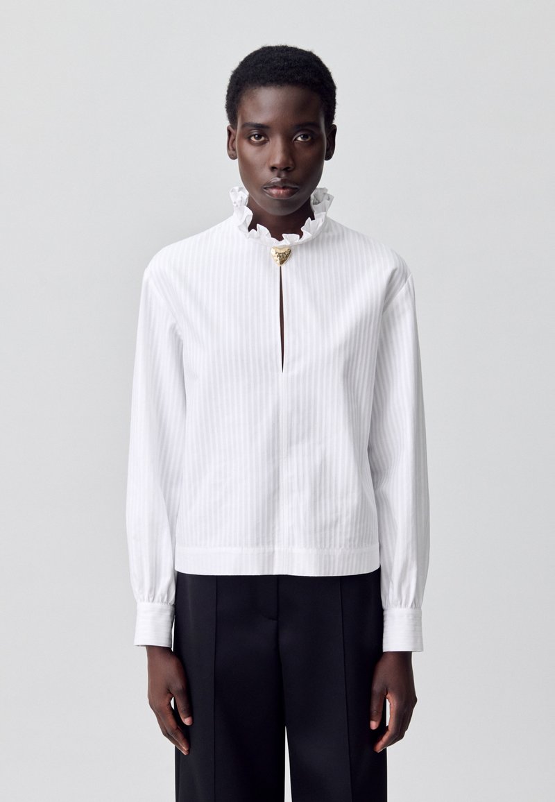 Modelo con una blusa blanca a rayas con un cuello con volantes y un broche dorado, combinada con pantalones negros, sobre un fondo neutro.