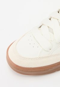 Hvit sneakers med overdel i semsket skinn og skinn, tykke kremfargede snøringer, perforert tå og en lys brun gummisåle.