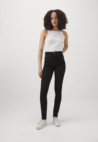 Fekete, magas derekú farmernadrág, testhez simuló fazonban, fehér ujjatlan felsővel, amelyen egy kis logó található. A modellen világos színű sportcipők vannak.