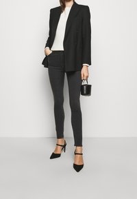 Blazer negro de doble botonadura sobre una blusa blanca, combinado con unos pantalones ajustados grises de cintura alta y tacones de ante negros, sosteniendo un pequeño bolso negro.