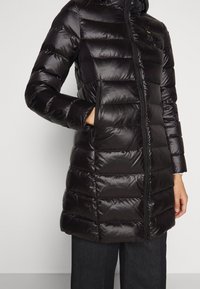 Czarna pikowana długa kurtka puchowa z przednim zamkiem błyskawicznym i bocznymi kieszeniami, noszona na ciemnych spodniach, ręka częściowo w kieszeni.