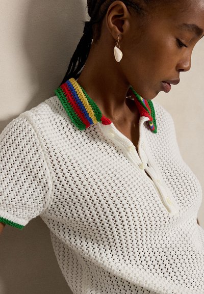 Γυναίκα που φοράει λευκό πλεκτό πόλο πουκάμισο με πολύχρωμο ριγέ γιακά και σκουλαρίκι σε σχήμα κοχυλιού, στραμμένη προφίλ μπροστά σε απλό φόντο.