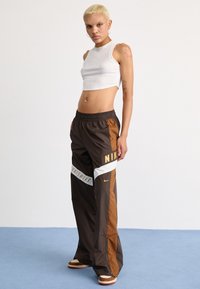 Pantaloni sportivi Nike di colore marrone con accenti arancioni, strisce bianche e grandi loghi. Presentano un bordo elastico in vita e dettagli con zip laterali.