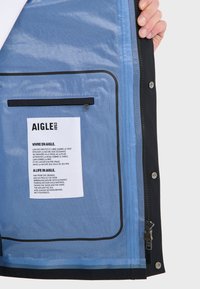 Sort inderside af sort jakke med blå foring, der har et mærke og en lynlås-lomme. Mærkets tekst er på fransk og engelsk, med accentuerede kanter.
